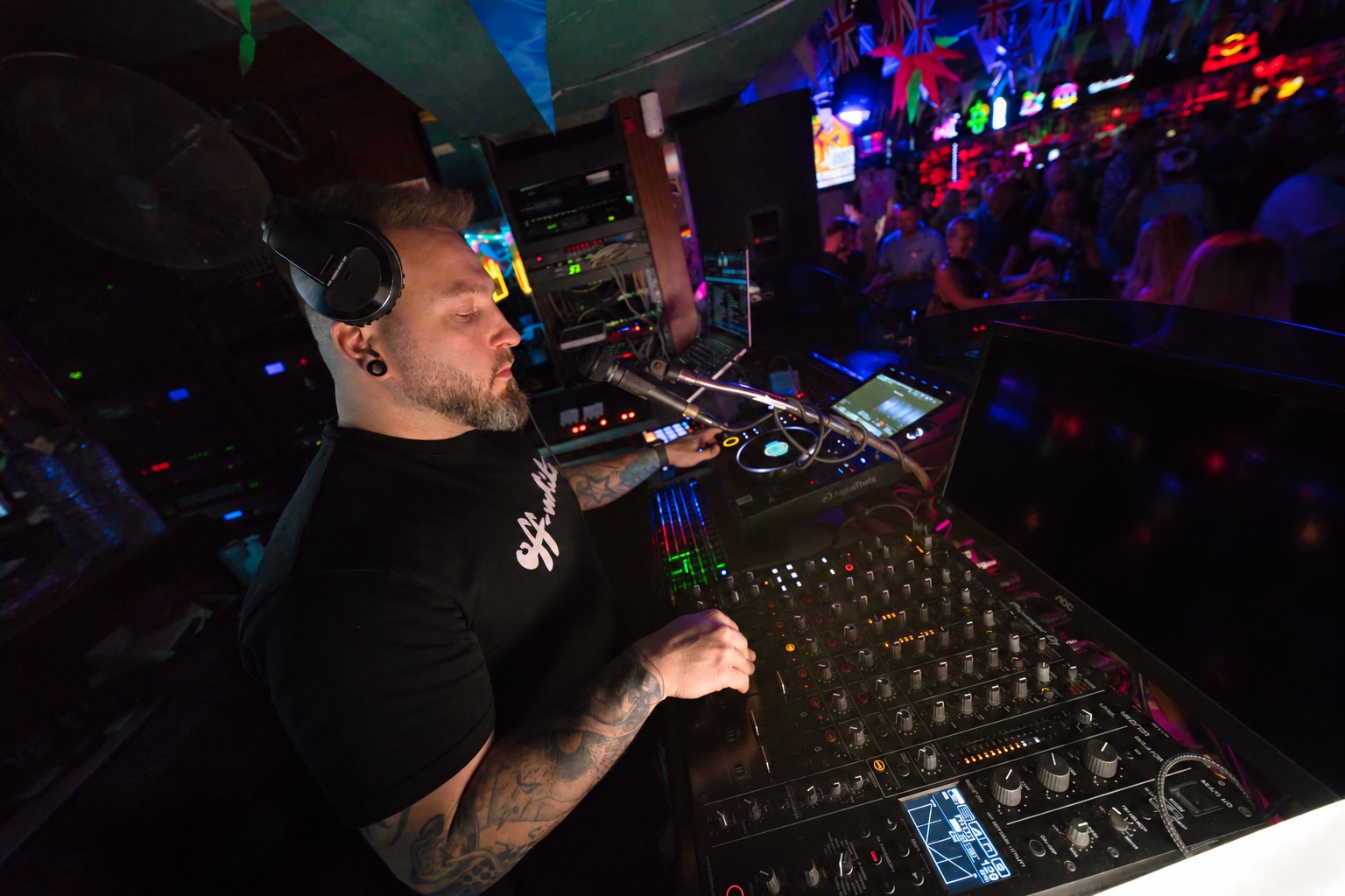 Ash Jenkins performing at a nightclub DJ booth with the crowd behind him